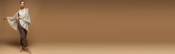 Longitud completa de la mujer afroamericana con estilo en chal de oro y pantalones de cuero posando en beige, bandera - foto de stock