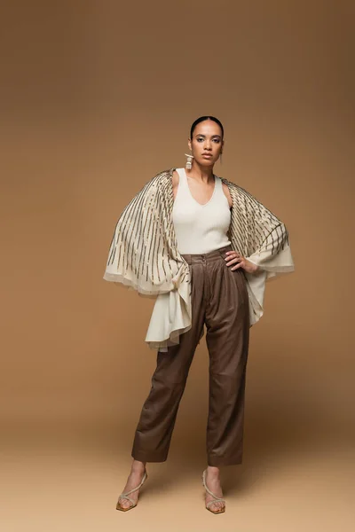 Full length of african american woman in golden shawl and leather pants posing with hands on hips on beige — Stock Photo