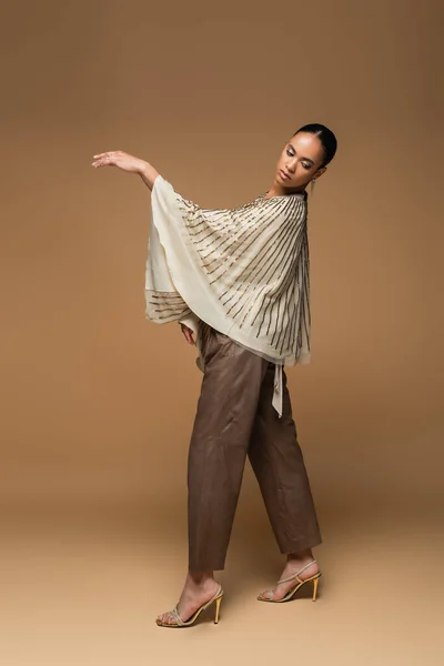 Full length of brunette african american model in golden shawl and leather pants posing on beige — Stock Photo