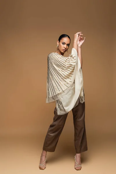 Full length of stylish african american model in golden jewelry and shawl posing on beige — Stock Photo