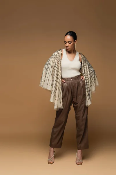 Piena lunghezza della donna afroamericana bruna in gioielli di lusso e scialle dorati in posa con le mani in tasca sul beige — Foto stock