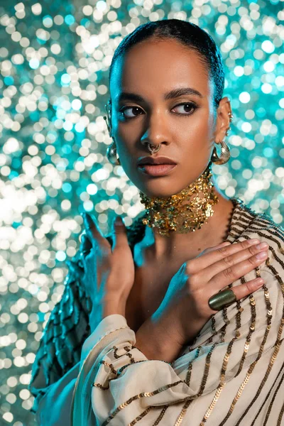 Проколота афроамериканська модель з золотою фольгою на шиї, позує схрещеними руками на блискучому синьому фоні — стокове фото