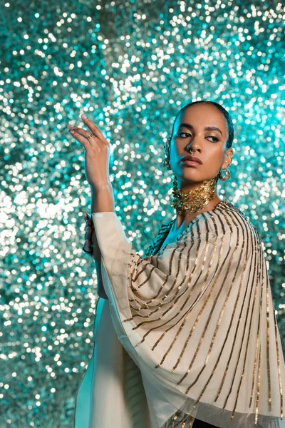 Modelo afroamericano perforado con lámina de oro en el cuello posando y mirando hacia otro lado sobre fondo azul brillante - foto de stock