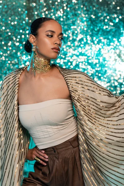 Проколота афроамериканська жінка з хусткою і золотом на шиї позує на блискучому синьому фоні — стокове фото