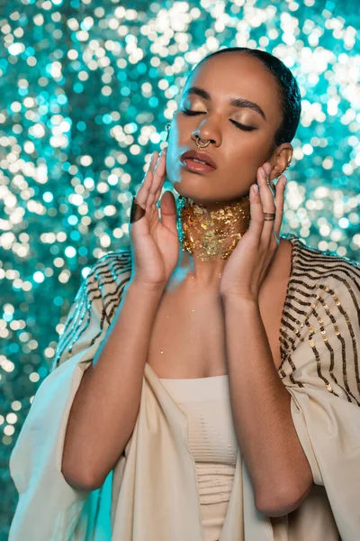 Brunette african american woman in shawl and gold on neck posing with closed eyes on sparkling blue background — Stock Photo