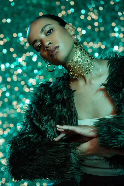 Young african american woman in faux fur jacket and gold on neck posing on sparkling turquoise background — Stock Photo