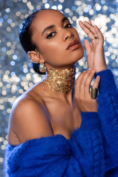 Mujer afroamericana de moda en chaqueta de piel sintética azul y accesorios dorados sobre fondo brillante - foto de stock