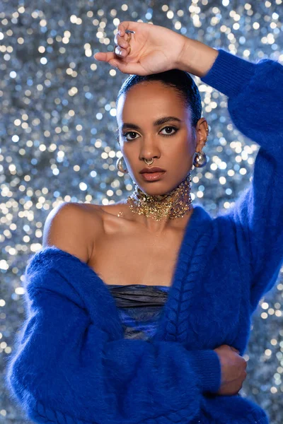 Modelo afroamericano de moda en suéter azul y accesorios dorados sobre fondo brillante - foto de stock