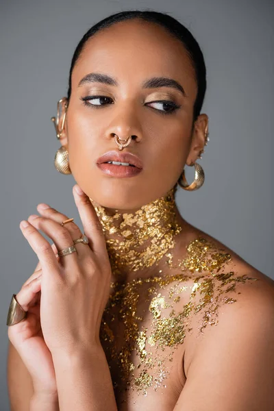 Joven mujer afroamericana con maquillaje dorado y lámina en el pecho mirando hacia otro lado aislado en gris - foto de stock