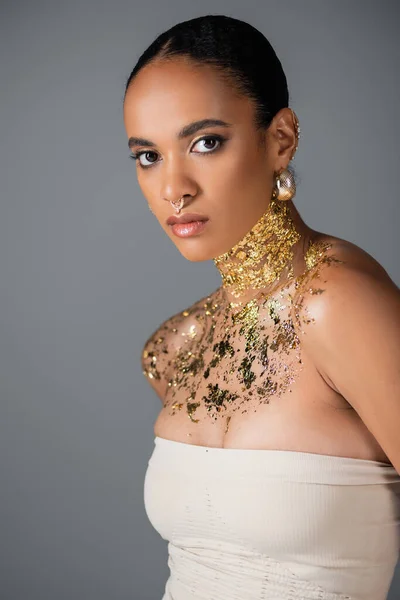 Femme afro-américaine à la mode avec percing et feuille d'or sur la poitrine en regardant la caméra isolée sur gris — Photo de stock