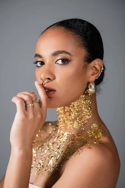 Afroamerikanerin mit goldenem Piercing und Folie auf der Brust berührt Lippen isoliert auf grau — Stockfoto