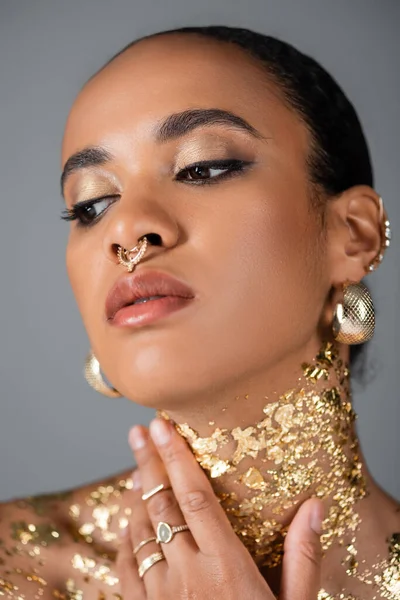 Portrait de jeune femme afro-américaine avec accessoires et percing touchant feuille d'or sur le cou isolé sur gris — Photo de stock