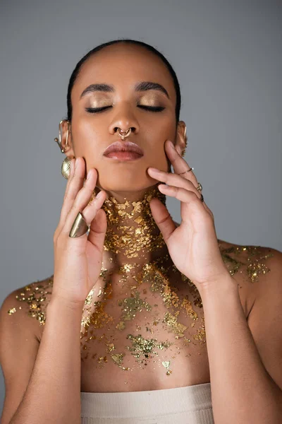 Mujer afroamericana bonita con maquillaje dorado y lámina en el pecho tocando mejillas aisladas en gris - foto de stock