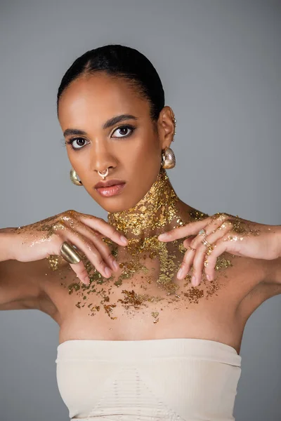 Fashionable african american woman with foil on body and golden accessories posing isolated on grey — Stock Photo