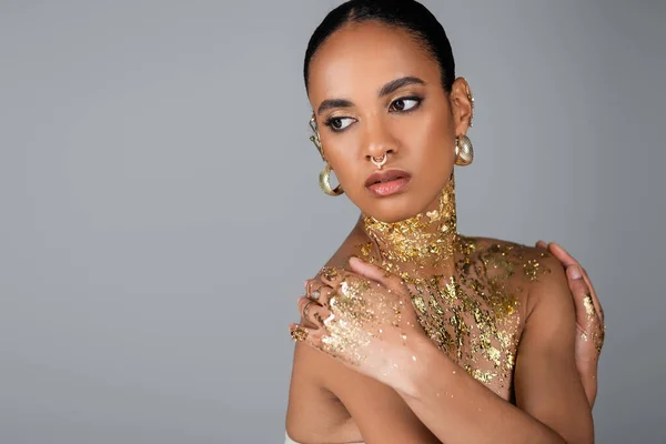 Stylish african american woman with golden foil on body touching shoulders isolated on grey — Stock Photo