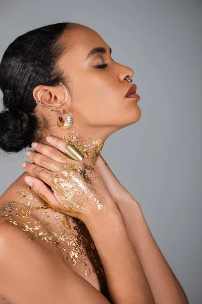 Vue latérale de la femme afro-américaine élégante avec feuille d'or sur les mains touchant le cou isolé sur gris — Photo de stock