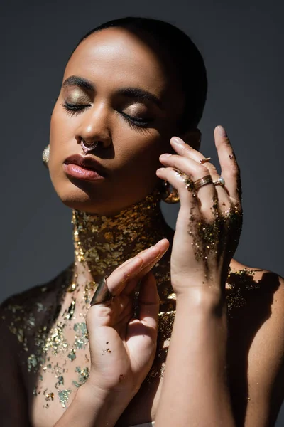 Donna afroamericana alla moda con piercing e lamina dorata sul corpo in posa in luce isolata sul grigio — Foto stock