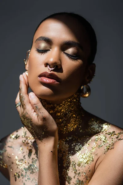 Портрет стильної афроамериканської моделі з золотим макіяжем і фольгою на тілі, що стоїть у світлі ізольовано на сірому — стокове фото