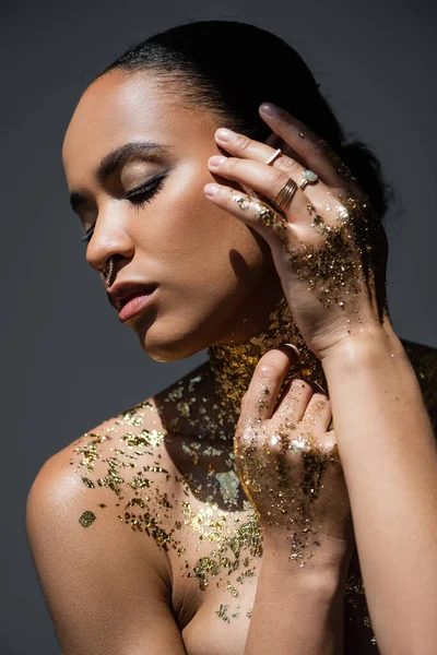 Young african american woman with golden foil on body touching face while posing in light isolated on grey — Stock Photo