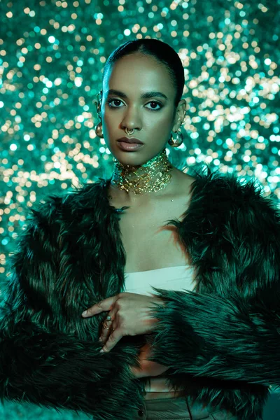 Donna afroamericana alla moda in pelliccia sintetica giacca e accessori guardando la fotocamera su sfondo scintillante — Foto stock