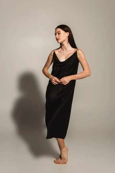 Босоногая модель с витилиго позирует в атласном платье на сером фоне с тенью — стоковое фото