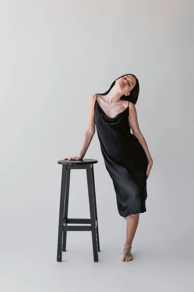 Femme sensuelle avec vitiligo en robe de soie touchant chaise sur fond gris — Photo de stock