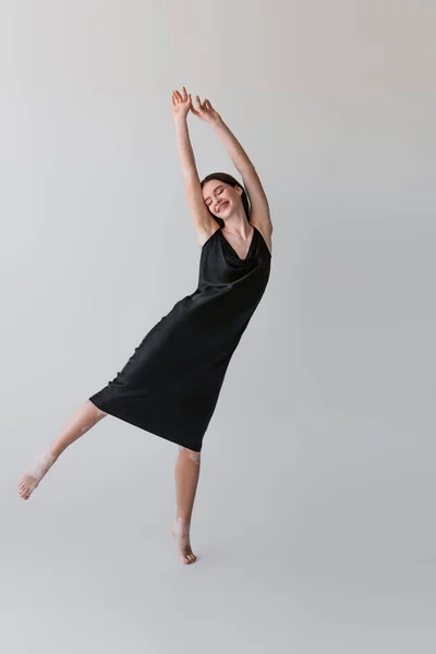 Longitud completa de mujer alegre con vitiligo levantando las manos mientras posa en vestido de seda sobre fondo gris - foto de stock