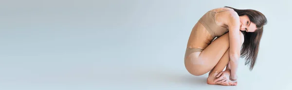 Modèle brune avec vitiligo assis en sous-vêtements beige avec soutien-gorge haut avec culotte sur fond gris, bannière — Photo de stock