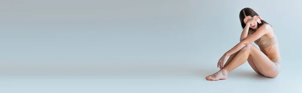 Повна довжина босоніж і молода жінка з вітіліго сидить у бежевій білизні і покриває обличчя на сірому, банер — стокове фото