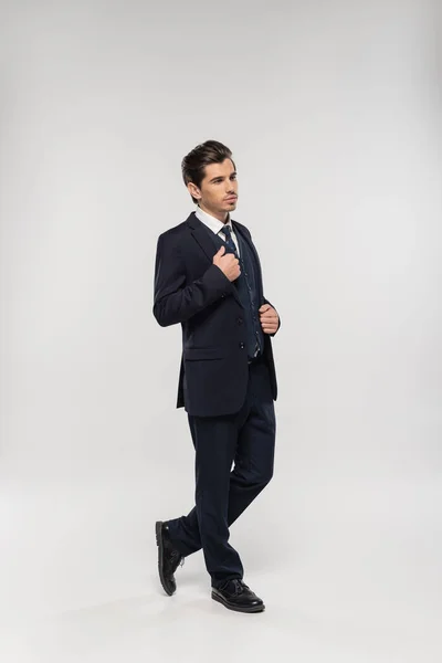 Повна довжина хорошого вигляду і молодого бізнесмена в костюмі коригування блейзера на сірому — Stock Photo