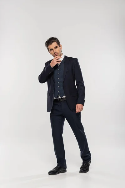 Piena lunghezza di successo e giovane uomo d'affari in giacca e cravatta toccando mento mentre posa sul grigio — Foto stock