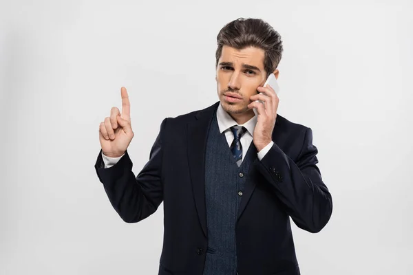 Young businessman in suit pointing up and talking on smartphone isolated on grey — Stock Photo