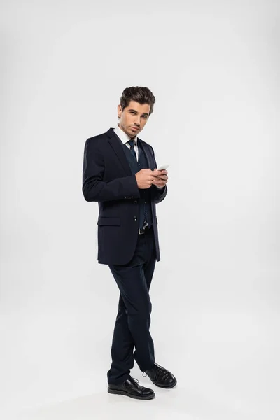 Повна довжина молодого і успішного бізнесмена в костюмі обміну повідомленнями на смартфоні на сірому — стокове фото
