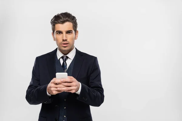 Aturdido y joven hombre de negocios en traje de mensajería en el teléfono inteligente aislado en gris - foto de stock