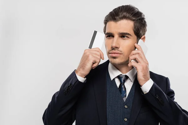 Jovem empresário em terno falando no smartphone e segurando cartão de crédito isolado no cinza — Fotografia de Stock