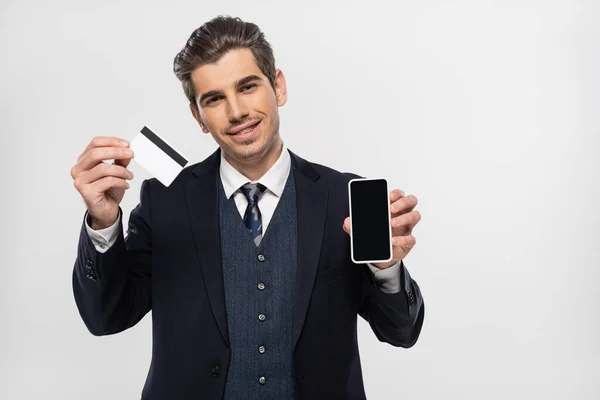 Alegre hombre de negocios con tirantes sosteniendo smartphone con pantalla en blanco y tarjeta de crédito aislada en gris - foto de stock