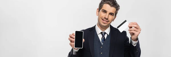 Cheerful businessman in braces holding smartphone with blank screen and credit card isolated on grey, banner — Stock Photo