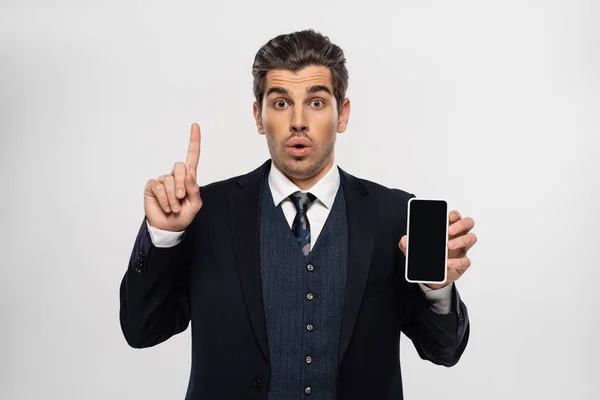 Sorprendido hombre de negocios en traje sosteniendo teléfono inteligente con pantalla en blanco y apuntando hacia arriba aislado en gris - foto de stock