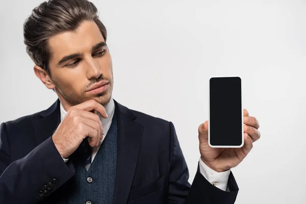 Вдумчивый бизнесмен в костюме смотрит на смартфон с пустым экраном, изолированным на сером — стоковое фото