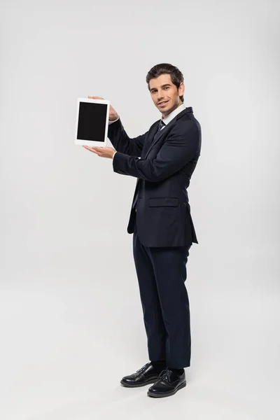 Повна довжина веселого бізнесмена в костюмі тримає цифровий планшет з порожнім екраном на сірому — стокове фото