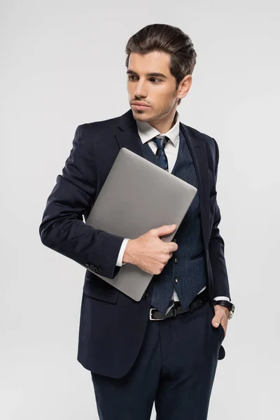 Joven hombre de negocios en traje que sostiene el ordenador portátil y de pie con la mano en el bolsillo aislado en gris - foto de stock