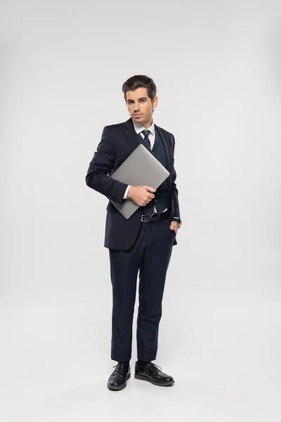 Full length of successful businessman in suit holding laptop and standing with hand in pocket on grey — Stock Photo