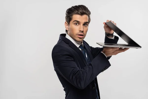 Приголомшений бізнесмен в костюмі дивиться на камеру і тримає ноутбук ізольований на сірому — стокове фото