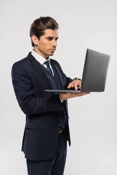 Hombre de negocios confiado en traje usando el ordenador portátil mientras que trabaja aislado remotamente en gris - foto de stock