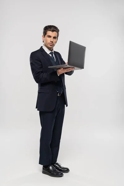 Full length of confident businessman in suit using laptop while working remotely on grey — Stock Photo