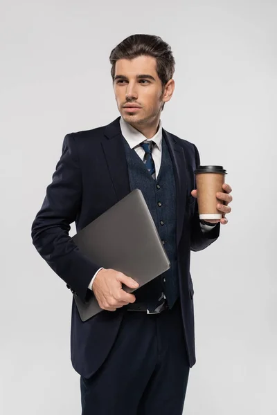 Confident businessman in suit holding paper cup and laptop isolated on grey — Stock Photo