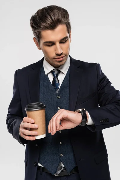 Joven empresario sosteniendo taza de papel con bebida para llevar mientras mira reloj de pulsera aislado en gris - foto de stock