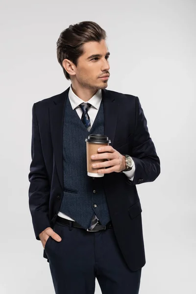Junger Geschäftsmann hält Pappbecher mit Imbissgetränk, während er mit der Hand in der Tasche isoliert auf grau steht — Stockfoto