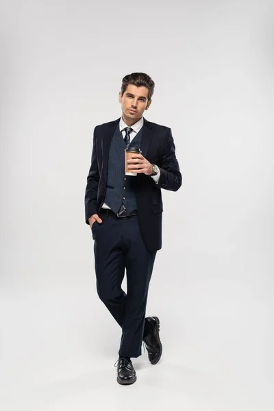 Полная длина молодого бизнесмена, держащего бумажную чашку с напитком на вынос, стоя с рукой в кармане на сером — стоковое фото