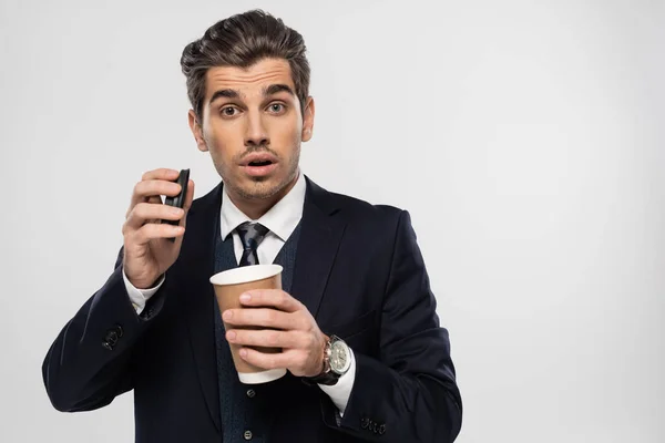 Sorprendido y soñoliento hombre de negocios sosteniendo taza de papel con bebida para llevar aislado en gris - foto de stock